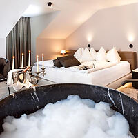 Luxurious hotel room with a round bathtub filled with foam, elegant candles, and a plush bed with a soft headboard in the background